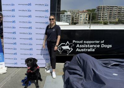 Smiling woman and black labrador assistance dog in front of Celestial V70 yacht which has Assistance Dogs Australia logo on its starboard side