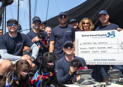 Two adult and two puppy labradors surrounded by smiling crew. Giant cheque being presented to female representative for Assistance Dogs Australia by Sam Haynes, owner of Sydney Animal Hospitals.