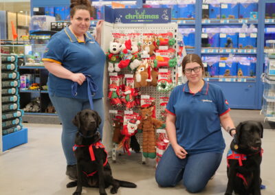 Two female Petstock employees pose with a black labrador assistance dog in front of their Christmas merchandise