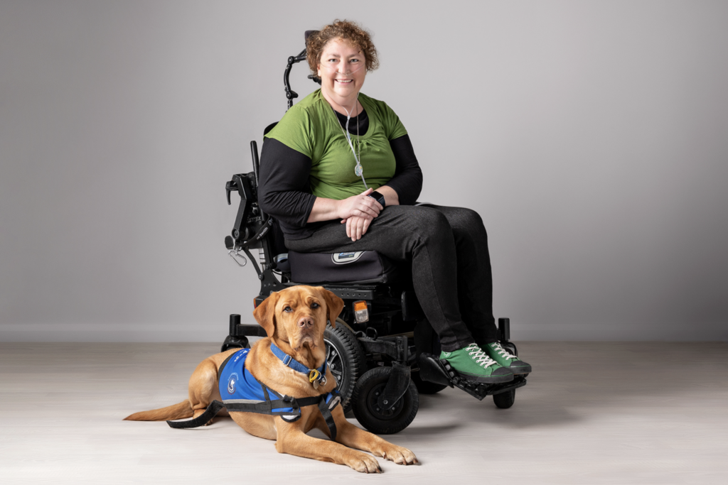 Smiling woman sitting in wheelchair next to her Assistance Dog lying on the ground beside her.